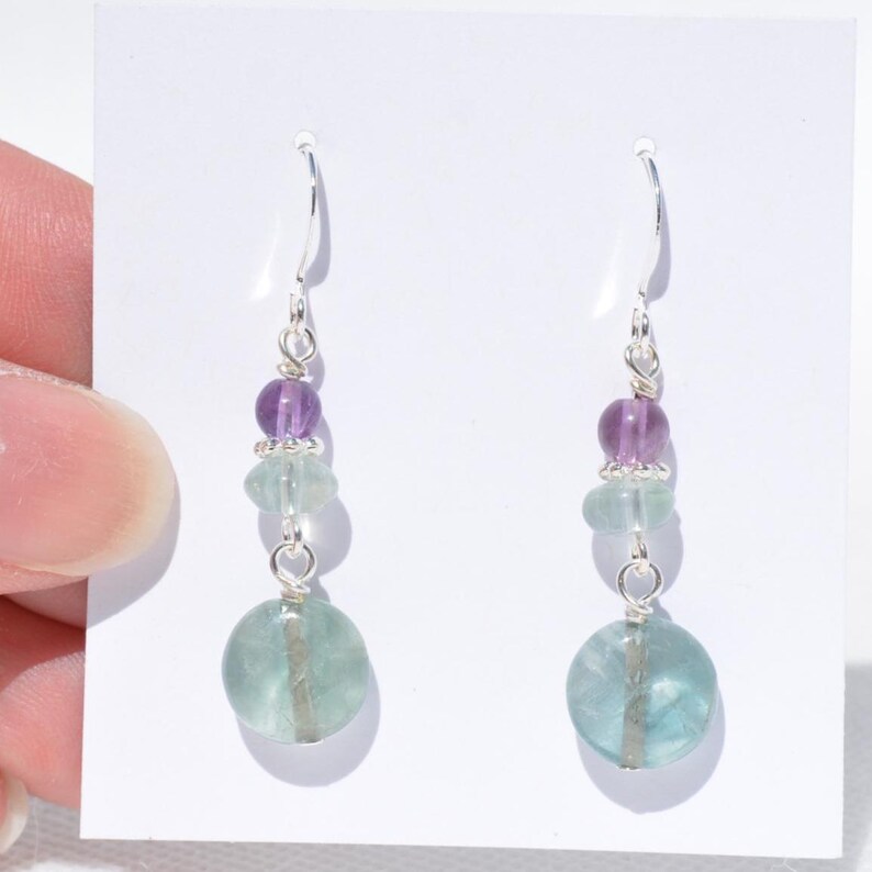 May include: Dangle earrings with purple and green beads. The earrings have silver hooks and wire, with a large, round, light green bead at the base. The beads are strung together, creating a layered effect. The earrings are designed to hang.