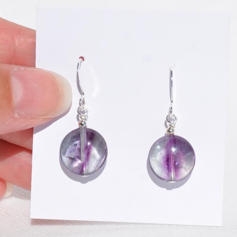 May include: A pair of dangle earrings with round, translucent purple and grey fluorite beads. The beads are suspended from silver-coloured ear wires. The earrings are displayed on a white background.