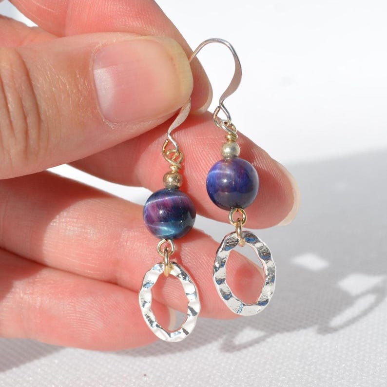 Op de afbeelding: Hangende oorbellen met ronde, dieppaarse en blauwe kattenoogkralen, geaccentueerd door kleine goudkleurige metalen bolletjes. Elke oorbel eindigt in een getextureerde, gehamerde zilverkleurige ovale ring die aan een eenvoudige metalen haak hangt.
