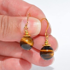 Puede incluir: Un par de pendientes colgantes con piedras preciosas de ojo de tigre redondas y pulidas, con bandas de color marr&oacute;n dorado. Cada piedra est&aacute; suspendida de un gancho de metal dorado y decorada con una peque&ntilde;a cuenta dorada en la parte superior.