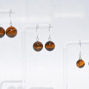 Puede incluir: Tres pares de pendientes colgantes con piedras preciosas esf&eacute;ricas de ojo de tigre suspendidas de ganchos de metal plateado. Cada par muestra las caracter&iacute;sticas bandas chatoyantes de color marr&oacute;n dorado, con el par de la derecha incluyendo peque&ntilde;as tapas decorativas plateadas sobre las esferas.