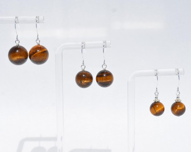 Puede incluir: Tres pares de pendientes colgantes con piedras preciosas esf&eacute;ricas de ojo de tigre suspendidas de ganchos de metal plateado. Cada par muestra las caracter&iacute;sticas bandas chatoyantes de color marr&oacute;n dorado, con el par de la derecha incluyendo peque&ntilde;as tapas decorativas plateadas sobre las esferas.