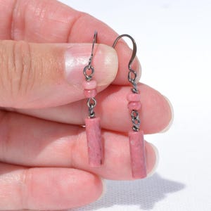 May include: A pair of drop earrings featuring dark gunmetal hooks and chains. Each earring is accented with two small, round, dusty pink beads followed by a single, cylindrical, dusty pink stone bead at the base, creating a simple, linear design.