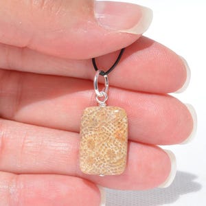 Op de afbeelding: Een rechthoekige hanger met een beige en zandkleurige gefossiliseerde koraalsteen met een gedetailleerde honingraatstructuur. De steen hangt aan een zilverkleurige metalen ring die is bevestigd aan een dun zwart koord, met zichtbare natuurlijke patronen.
