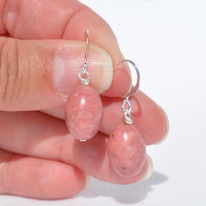 Peut inclure: Une paire de boucles d'oreilles pendantes compos&eacute;es de pierres de rhodonite rose de forme ovale lisse, suspendues &agrave; des crochets en m&eacute;tal argent&eacute;. Chaque pierre pr&eacute;sente une texture marbr&eacute;e avec de fines inclusions sombres.