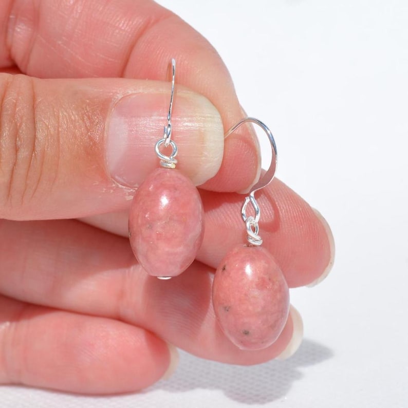 Peut inclure: Une paire de boucles d'oreilles pendantes compos&eacute;es de pierres de rhodonite rose de forme ovale lisse, suspendues &agrave; des crochets en m&eacute;tal argent&eacute;. Chaque pierre pr&eacute;sente une texture marbr&eacute;e avec de fines inclusions sombres.