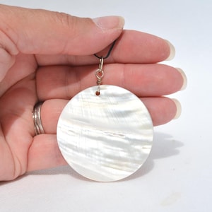May include: A round, white pendant with a pearlescent sheen. The pendant is suspended from a black cord and attached with a silver-colored metal loop. The pendant is held in a hand against a white background.