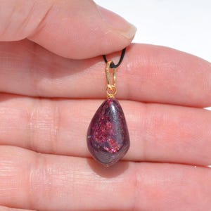 May include: A teardrop-shaped pendant with a deep burgundy hue, suspended from a black cord and a gold-colored bail. The pendant's surface has a shimmering, textured appearance, catching the light. The jewelry piece is held in a hand.