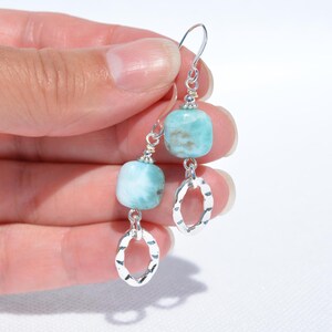 May include: A pair of dangle earrings featuring light blue, square-cut gemstones and silver-colored, hammered oval charms. The earrings have a simple hook design and are displayed against a white background.