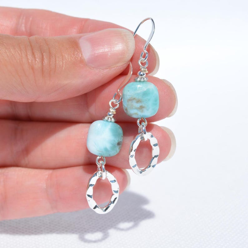 May include: A pair of dangle earrings featuring light blue, square-cut gemstones and silver-colored, hammered oval charms. The earrings have a simple hook design and are displayed against a white background.