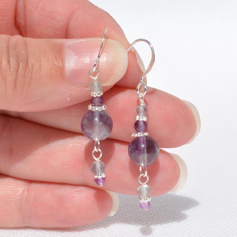 May include: Dangle earrings featuring a combination of purple and clear beads. The earrings have a silver hook and are composed of various sizes of round and oval beads. The earrings are a light purple color.