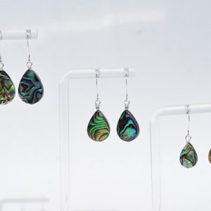 May include: Three pairs of teardrop-shaped earrings with iridescent abalone shells. The earrings hang from silver-colored hooks. The abalone shells display a mix of green, blue, and brown colors, with unique patterns. The earrings are displayed on white stands.