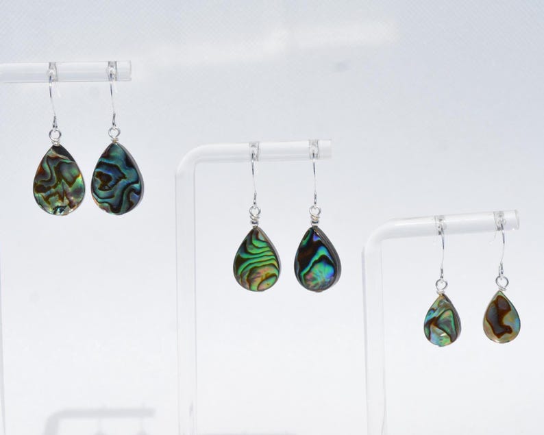 May include: Three pairs of teardrop-shaped earrings with iridescent abalone shells. The earrings hang from silver-colored hooks. The abalone shells display a mix of green, blue, and brown colors, with unique patterns. The earrings are displayed on white stands.