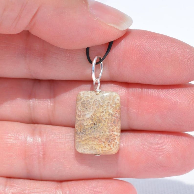Op de afbeelding: Een rechthoekige hanger met een getextureerd, beige en lichtbruin oppervlak van versteende steen met een poreus uiterlijk. De steen is bevestigd aan een zilverkleurige metalen ring die aan een dun zwart koord is bevestigd.