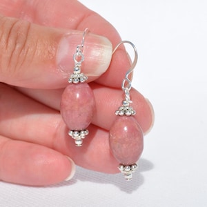 Peut inclure: Une paire de boucles d'oreilles pendantes compos&eacute;es de perles ovales et lisses en rhodonite rose. Chaque perle est orn&eacute;e de petites coupelles d&eacute;coratives en m&eacute;tal argent&eacute; en haut et en bas, suspendues &agrave; des crochets d'oreilles simples.