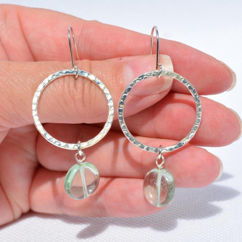 May include: Silver hoop earrings with a textured finish. Each earring has a clear, oval-shaped, pale green gemstone pendant. The earrings are shown against a light background.