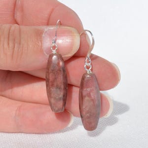 May include: A pair of drop earrings featuring elongated, cylindrical beads in a mottled dusty rose and grey stone pattern. Each bead is suspended from a simple silver-toned metal hook wire, creating a clean and straightforward design for these dangle earrings.