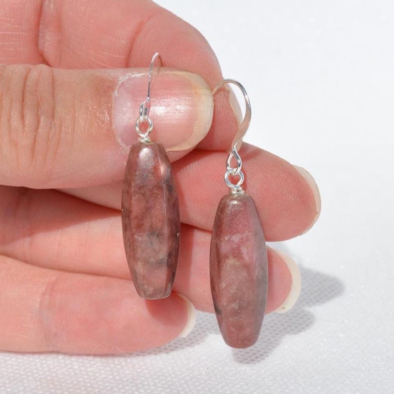 May include: A pair of drop earrings featuring elongated, cylindrical beads in a mottled dusty rose and grey stone pattern. Each bead is suspended from a simple silver-toned metal hook wire, creating a clean and straightforward design for these dangle earrings.