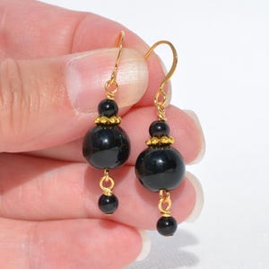 May include: A pair of dangle earrings featuring two smooth, round black beads of different sizes, separated and accented by small, textured gold-tone metal spacers and connected by gold-tone wire hooks for pierced ears.