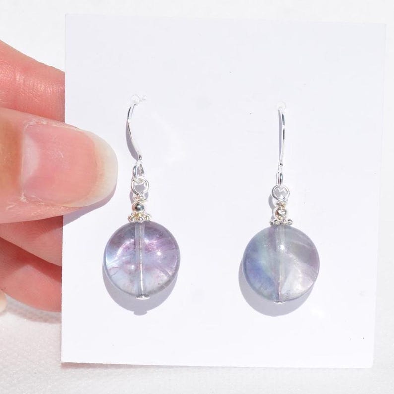 May include: A pair of earrings with round, translucent, purple and blue fluorite beads. The beads hang from silver-coloured hooks with small decorative elements. The earrings are shown against a white background.