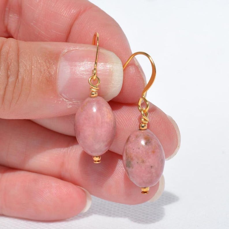Peut inclure: Une paire de boucles d'oreilles pendantes compos&eacute;es de perles ovales et lisses en rhodonite rose, suspendues &agrave; des crochets en m&eacute;tal dor&eacute;. Chaque perle pr&eacute;sente des motifs naturels marbr&eacute;s dans des tons vieux rose et rose p&acirc;le, fix&eacute;es par des fils dor&eacute;s.