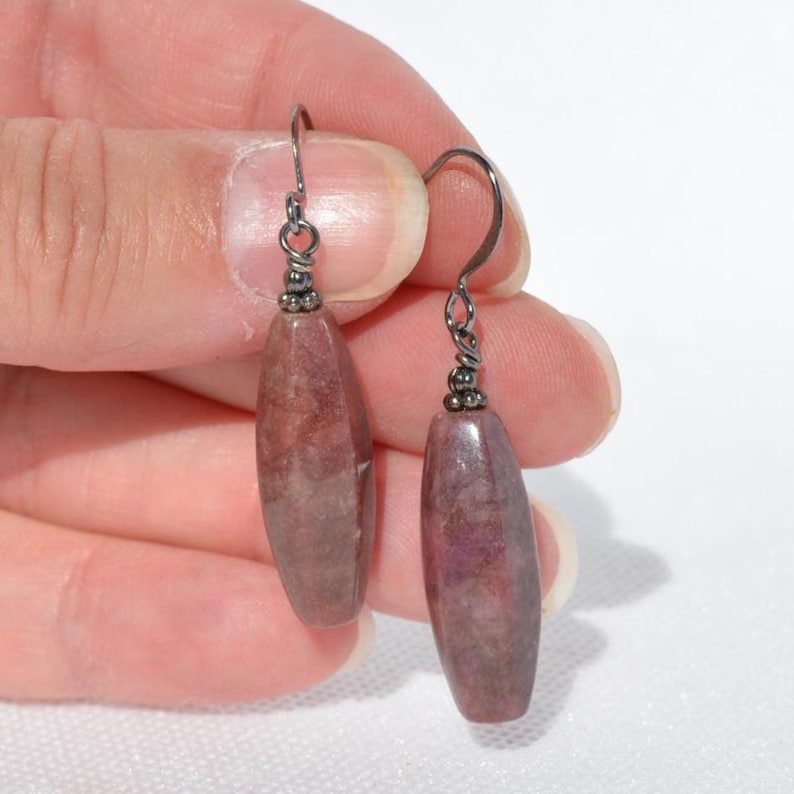 May include: A pair of drop earrings featuring elongated, faceted, dusty rose-coloured stone beads suspended from dark metal hooks. Each bead has a smooth, polished surface with subtle internal variations in colour and texture, attached to a small metal loop.