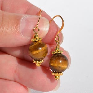 Puede incluir: Un par de pendientes colgantes con piedras preciosas de ojo de tigre esf&eacute;ricas de brillo marr&oacute;n dorado. Cada cuenta est&aacute; acentuada por peque&ntilde;as cuentas de metal dorado en la parte superior e inferior, suspendidas de ganchos dorados curvos.