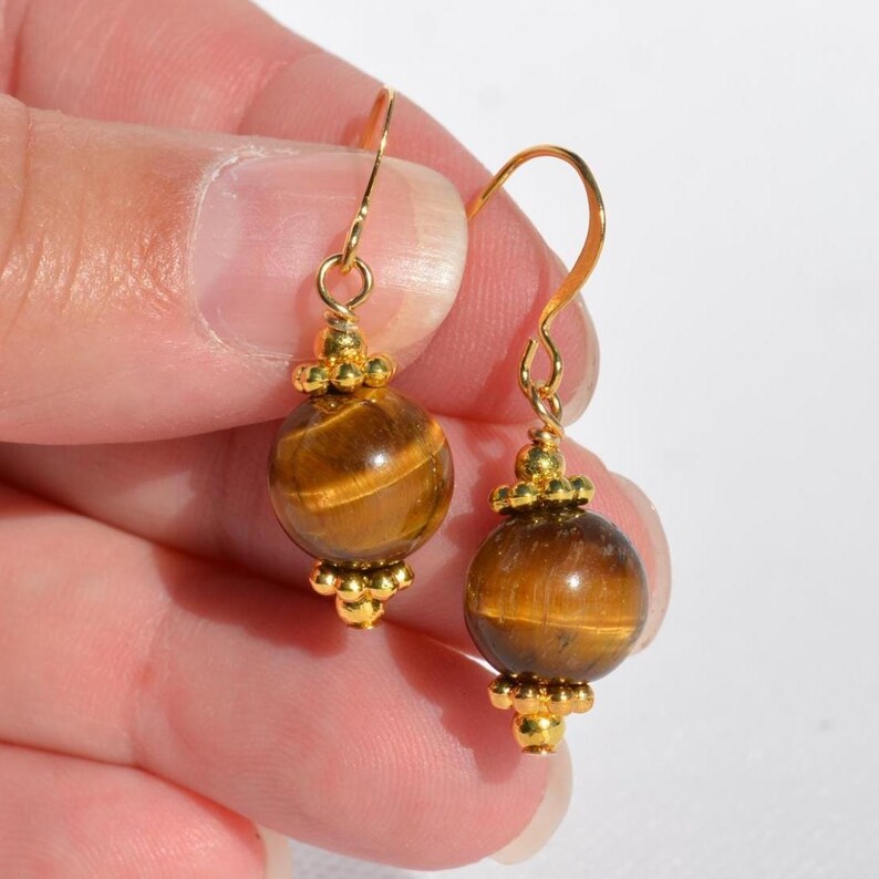 Puede incluir: Un par de pendientes colgantes con piedras preciosas de ojo de tigre esf&eacute;ricas de brillo marr&oacute;n dorado. Cada cuenta est&aacute; acentuada por peque&ntilde;as cuentas de metal dorado en la parte superior e inferior, suspendidas de ganchos dorados curvos.