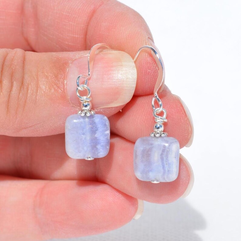 May include: A pair of silver-toned drop earrings featuring square, light blue gemstones. The earrings have a simple design with a hook closure and small decorative silver accents. The gemstones have a subtle, striped pattern.