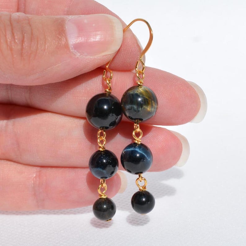 Op de afbeelding: Hangende oorbellen met drie trapsgewijze ronde blauwe tijgeroog edelsteenkralen opgehangen aan een goudkleurige metalen draad. De kralen vertonen een donkerblauwe en zwarte chatoyante glans, verbonden door kleine goudkleurige ringetjes.