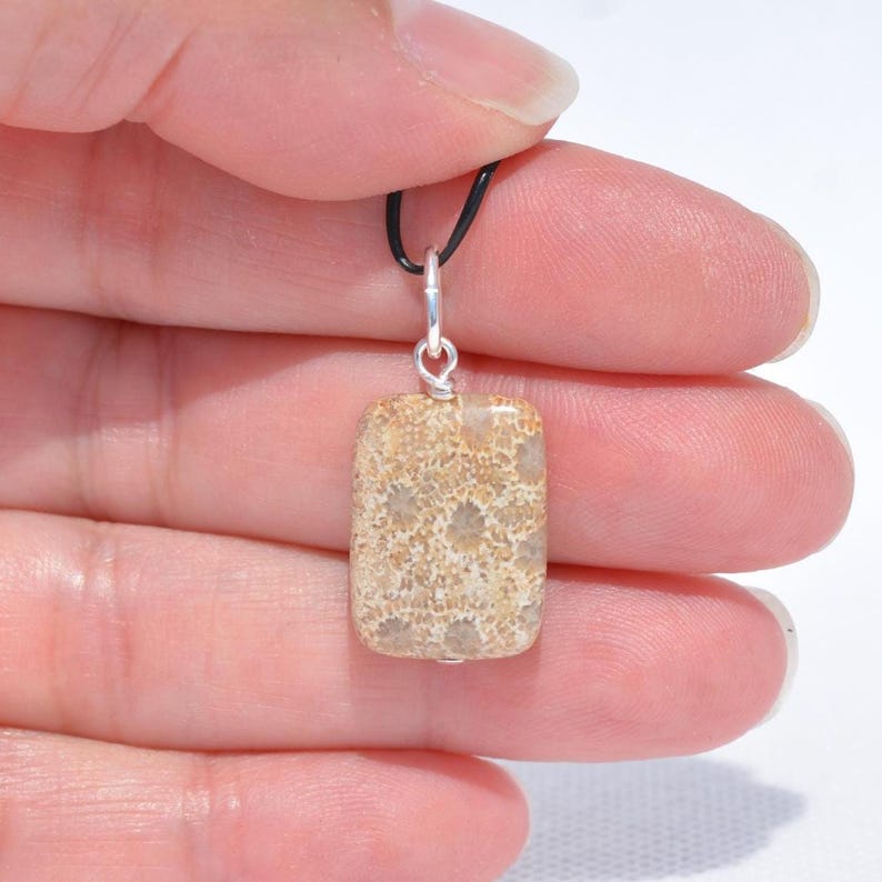 Op de afbeelding: Een rechthoekige hanger met een beige en zandkleurige gefossiliseerde koraalsteen met een natuurlijk gespikkeld patroon. De steen is bevestigd aan een zilverkleurig metalen oogje en hangt aan een dun zwart koord, met een gladde, gepolijste afwerking.