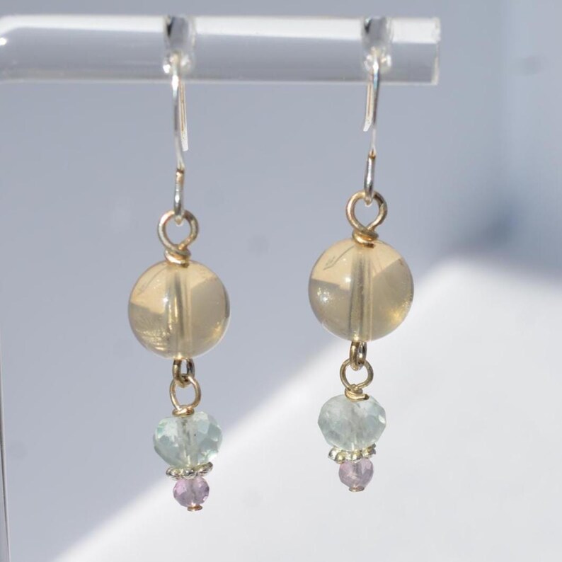 May include: A pair of dangle earrings with silver hooks. Each earring features a large, round, translucent, light-yellow bead, followed by a smaller, light-blue bead and a tiny, round, pink bead. The earrings are suspended from a clear display stand.