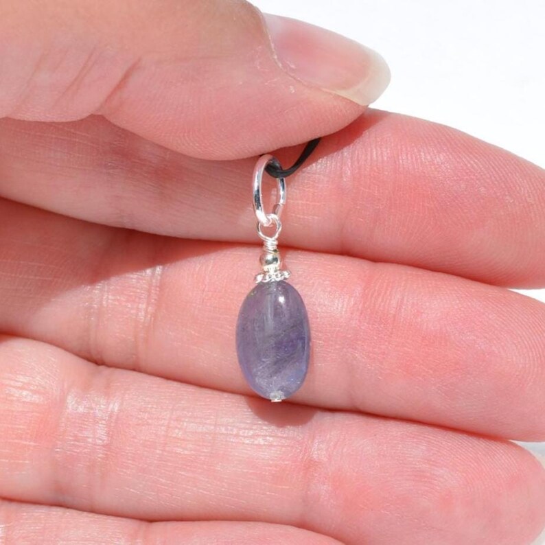 May include: A small, oval-shaped, lavender-colored gemstone pendant with a silver-colored bail. The pendant is suspended from a black cord. The gemstone is smooth and polished, and the bail has decorative detailing.