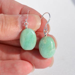 May include: A pair of drop earrings featuring smooth, oval-shaped light green stones suspended from silver-toned metal hooks. The stones exhibit natural veining and a polished finish, with each earring attached to a simple wire loop mechanism.