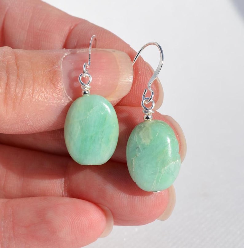 May include: A pair of drop earrings featuring smooth, oval-shaped light green stones suspended from silver-toned metal hooks. The stones exhibit natural veining and a polished finish, with each earring attached to a simple wire loop mechanism.