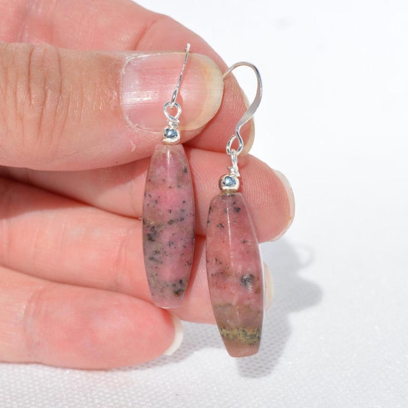 May include: A pair of drop earrings featuring elongated, faceted rhodonite beads in shades of dusty pink and black. The stones are suspended from silver-toned metal hooks, each accented with a small, round silver bead at the top of the stone.