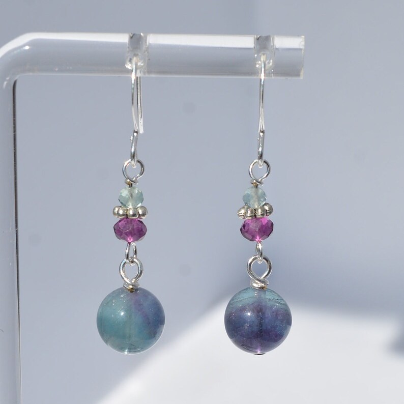May include: A pair of dangle earrings with silver-tone hooks. Each earring features a large, round, blue-green and purple fluorite bead, accented with smaller pink and green beads. The earrings are displayed against a light background.
