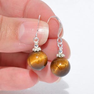 Puede incluir: Un par de pendientes colgantes con piedras preciosas de ojo de tigre esf&eacute;ricas con un brillo dorado y marr&oacute;n. Cada cuenta est&aacute; suspendida de un gancho de metal plateado y decorada con una peque&ntilde;a tapa de cuenta decorativa de color plateado.