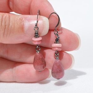 Peut inclure: Une paire de boucles d'oreilles pendantes avec des crochets en m&eacute;tal gris fonc&eacute; et des attaches en fil m&eacute;tallique. Chaque boucle comporte une petite perle disque rose clair au-dessus d'un pendentif en pierre rose mouchet&eacute; en forme de goutte.