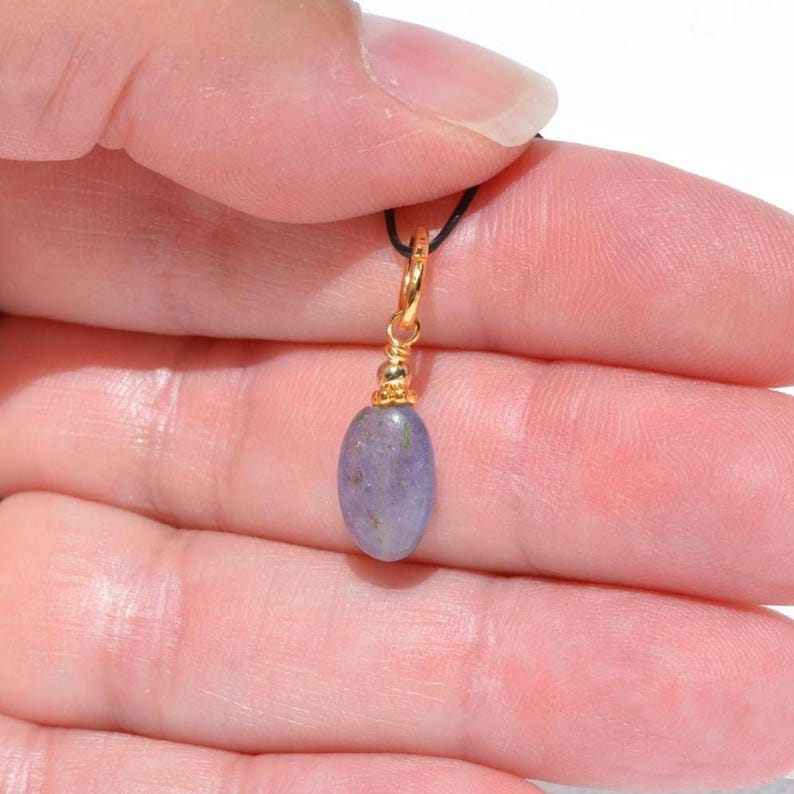 May include: A gold-colored pendant with a small, oval, lavender-colored stone. The pendant is suspended from a thin, black cord. The stone has a smooth, polished surface and is held in place by a decorative gold-colored setting.