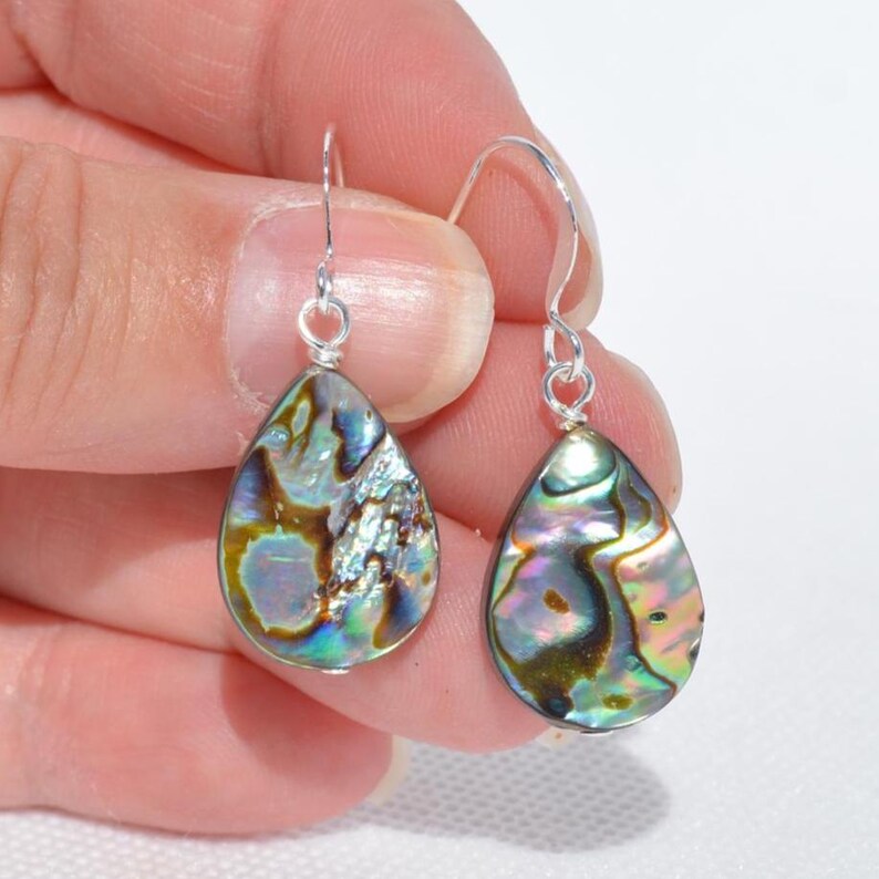 May include: A pair of teardrop-shaped earrings with iridescent abalone shells. The earrings have silver-colored hooks. The abalone shells display a mix of colors, including blues, greens, and browns, with a pearlescent sheen. The earrings are held in a hand.