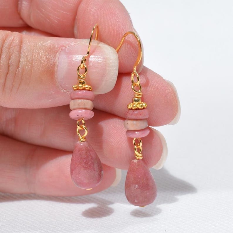 Peut inclure: Une paire de boucles d'oreilles pendantes avec des crochets et des &eacute;l&eacute;ments en m&eacute;tal dor&eacute;. Chaque boucle se compose de deux petites perles rondes rose poudr&eacute; surmontant un pendentif en pierre rose en forme de goutte, reli&eacute;s par des anneaux dor&eacute;s.