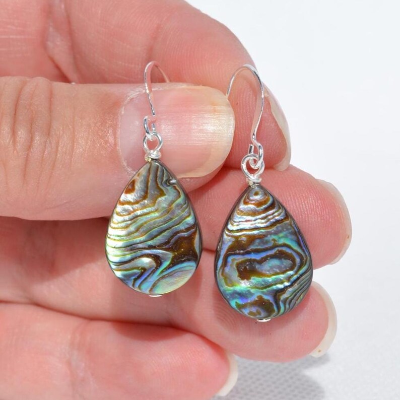 May include: A pair of teardrop-shaped earrings with iridescent abalone shells. The earrings have silver-colored hooks. The abalone shells display a mix of brown, blue, and green hues, with a pearlescent sheen. The earrings are held in a hand.