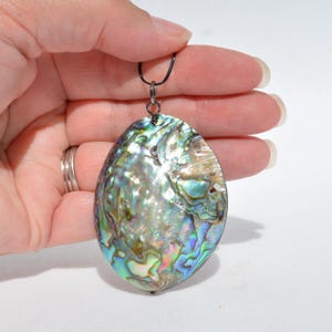 May include: Oval-shaped abalone shell pendant with iridescent colors of blue, green, and purple. The pendant is suspended from a black cord. The shell's surface has a natural, textured appearance, reflecting light.