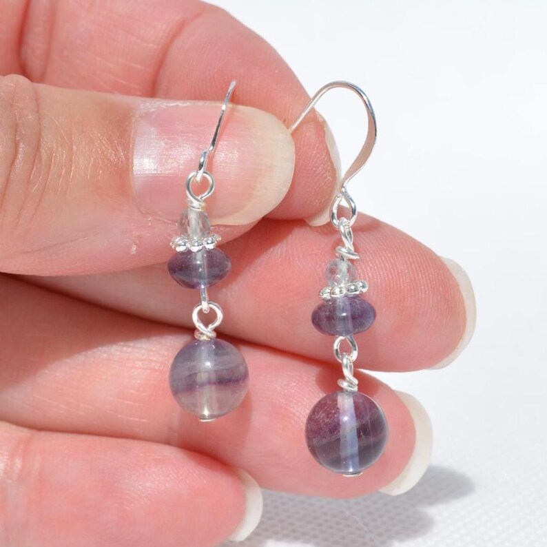 May include: Dangle earrings with a silver hook and wire design. Each earring features a clear bead cluster, a purple oval bead, and a large, round, purple and white striped bead. The earrings are held in a hand.
