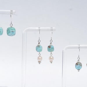 May include: Earrings with silver hooks and various designs. One pair features square, light blue beads. Another pair combines light blue beads with a pearl. The third pair has light blue beads with a silver accent. All earrings are displayed on white stands.