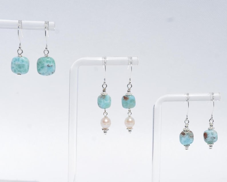 May include: Earrings with silver hooks and various designs. One pair features square, light blue beads. Another pair combines light blue beads with a pearl. The third pair has light blue beads with a silver accent. All earrings are displayed on white stands.