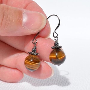 Puede incluir: Un par de pendientes colgantes con cuentas redondas de ojo de tigre pulido con reflejos marr&oacute;n dorado. Cada cuenta est&aacute; rematada con una peque&ntilde;a tapa decorativa de metal gris oscuro y suspendida de un gancho de metal oscuro a juego.