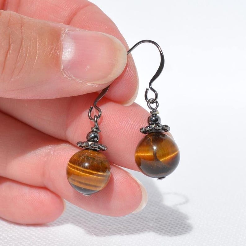 Puede incluir: Un par de pendientes colgantes con cuentas redondas de ojo de tigre pulido con reflejos marr&oacute;n dorado. Cada cuenta est&aacute; rematada con una peque&ntilde;a tapa decorativa de metal gris oscuro y suspendida de un gancho de metal oscuro a juego.