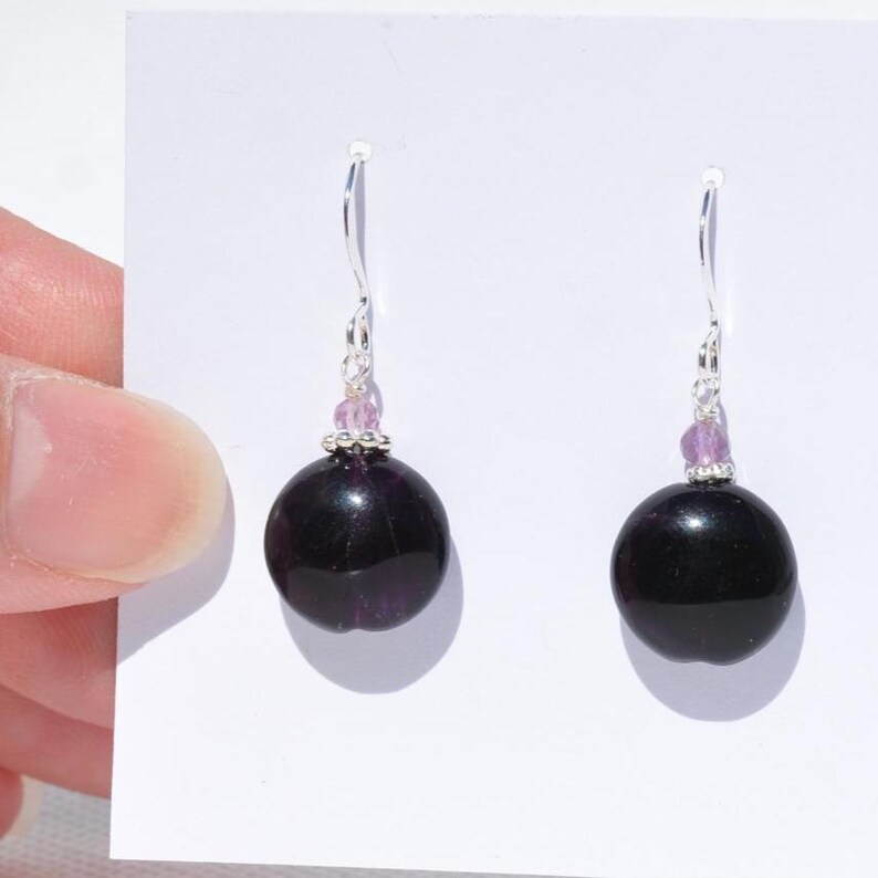 May include: A pair of drop earrings with large, dark purple, spherical beads. Each earring has a silver hook and is accented with small, light purple beads. The earrings are shown against a white background.